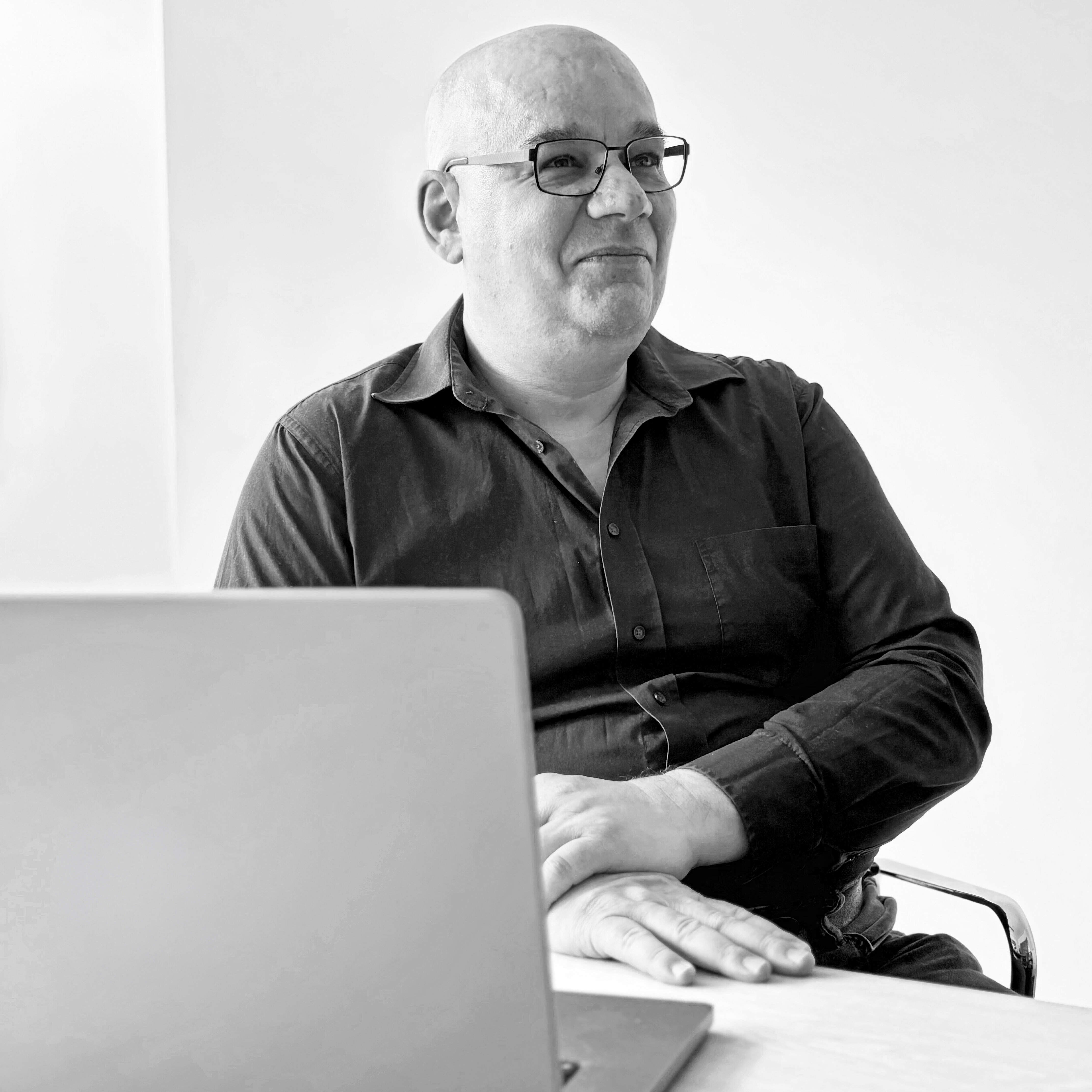Ein glatzköpfiger Mann mit Brille sitzt an einem Tisch hinter einem aufgeklappten Laptop und blickt leicht zur Seite. Die Aufnahme ist in Schwarz-Weiß gehalten und zeigt eine ruhige, konzentrierte Atmosphäre.