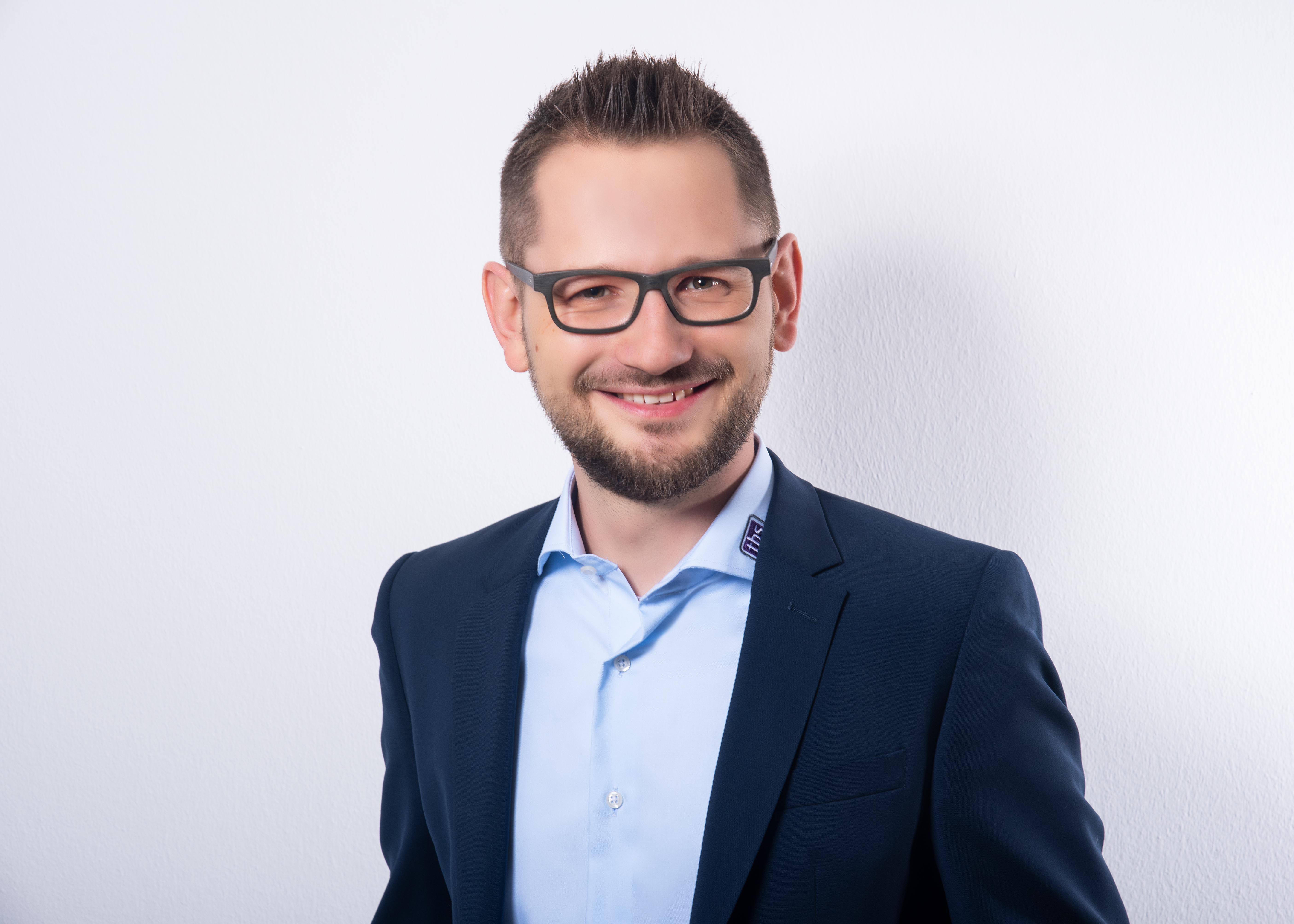 Mario Vogt, Geschäftsführer von tbs mit kurzen, gestylten Haaren, Brille und Bart lächelt in die Kamera; er trägt ein dunkelblaues Sakko über einem hellblauen Hemd. Der Hintergrund ist einfarbig weiß.