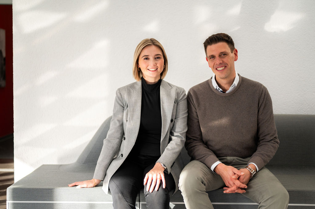 Zwei Personen sitzen nebeneinander auf einem grauen Sofa vor einer weißen Wand, beide lächeln in die Kamera. Die Frau trägt einen grauen Blazer und der Mann einen braunen Pullover über einem Hemd.