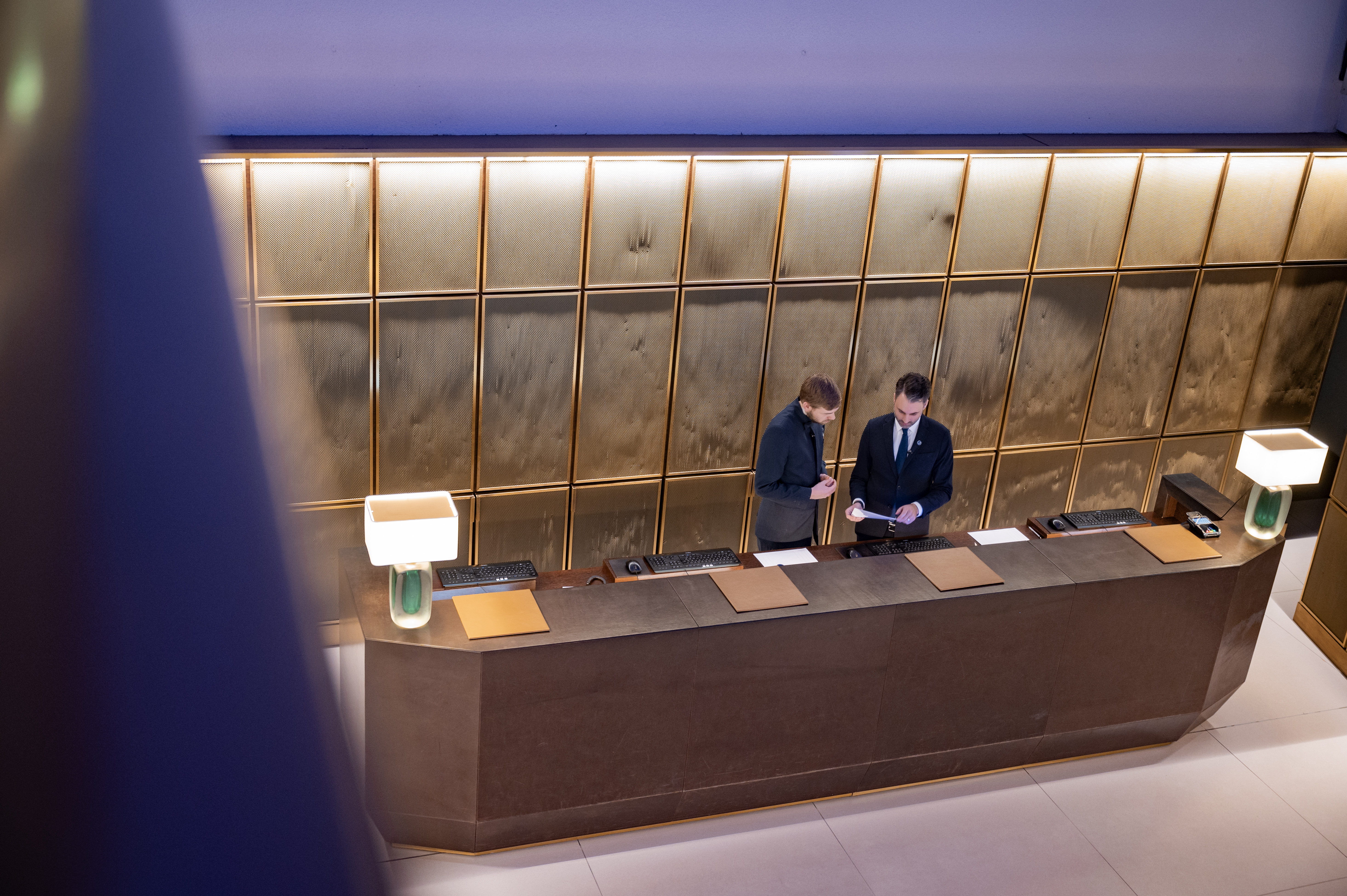 Zwei Männer in Anzügen stehen hinter einem elegant beleuchteten Empfangstresen und betrachten gemeinsam ein Dokument. Die Rückwand besteht aus goldfarben gerahmten Paneelen, auf dem Tresen stehen zwei moderne Tischlampen mit grünem Sockel.