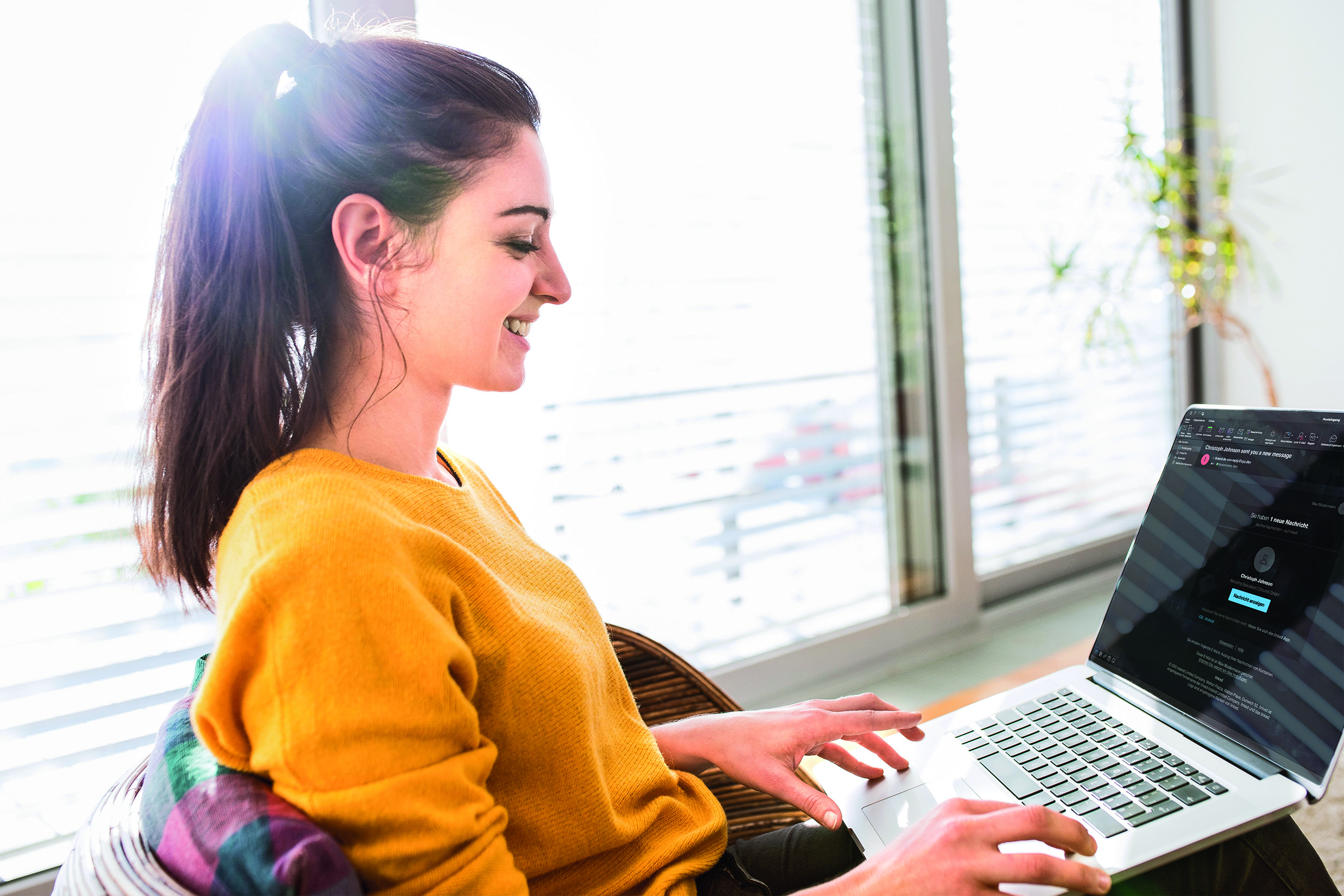 Eine junge Frau mit zusammengebundenen dunklen Haaren sitzt in einem hellen Raum am Fenster und arbeitet lächelnd an einem Laptop. Auf dem Bildschirm ist eine Messaging- oder E-Mail-Oberfläche mit einer neuen Nachricht und einem Profilnamen zu sehen, der Text ist jedoch nur teilweise lesbar.