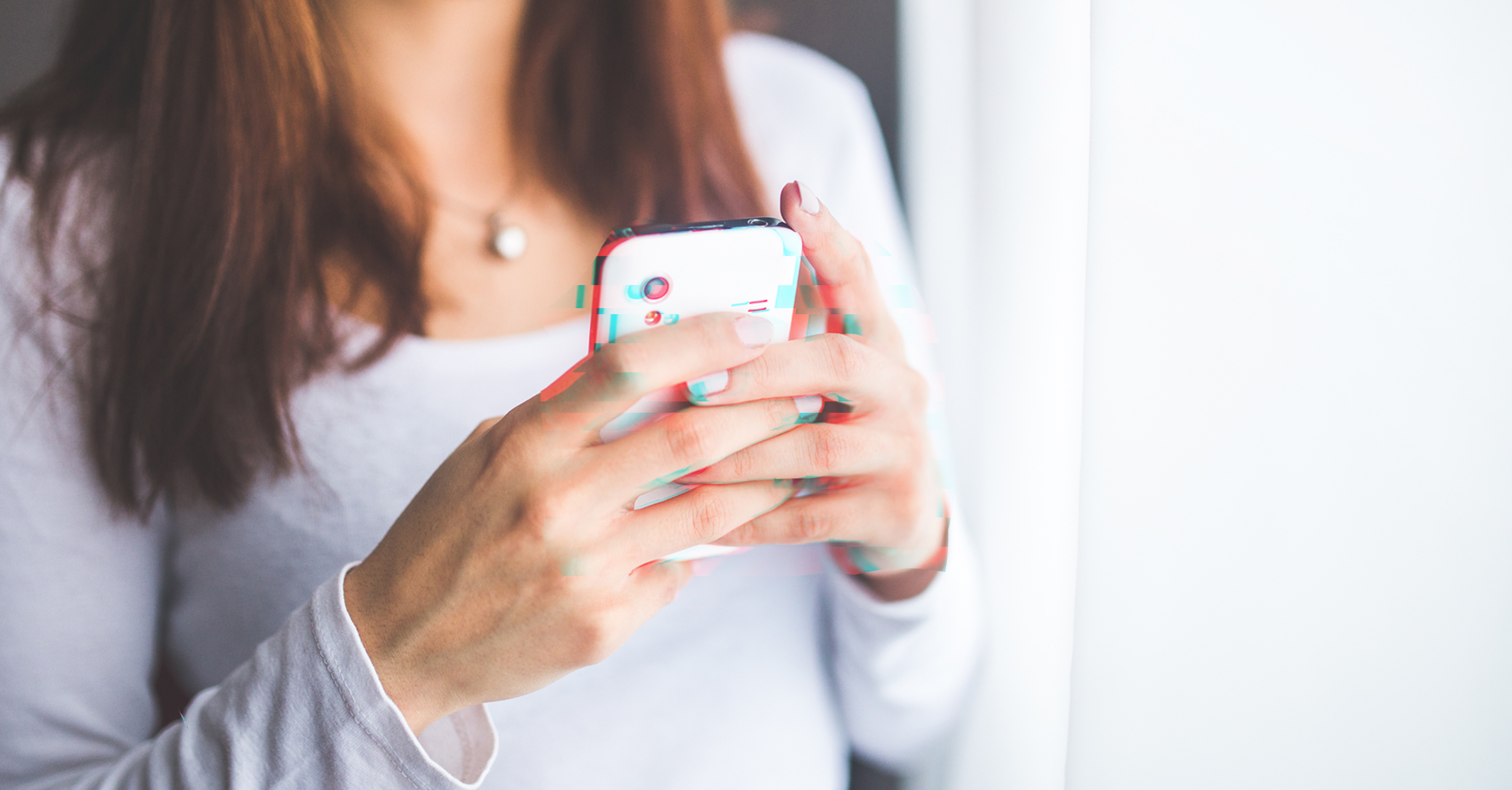 Eine Frau mit braunem schulterlangem haar und einem weißen Pullover, die in der Hand ein Smartphone hält.