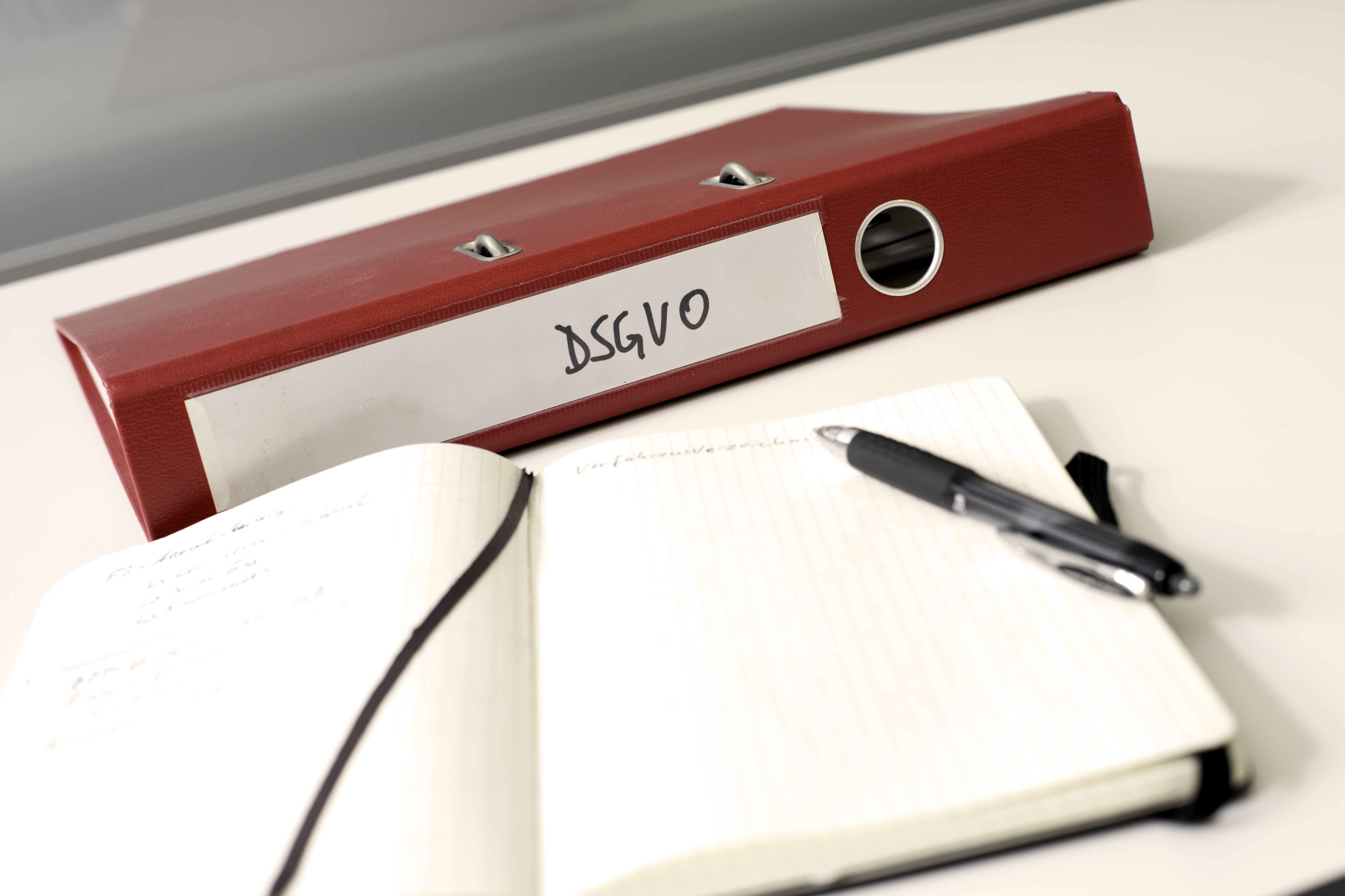 Ein roter Ordner mit der Aufschrift DSGVO. Davor ein aufgeschlagenes Notizbuch mit einem Stift. Die Seiten sind leer.